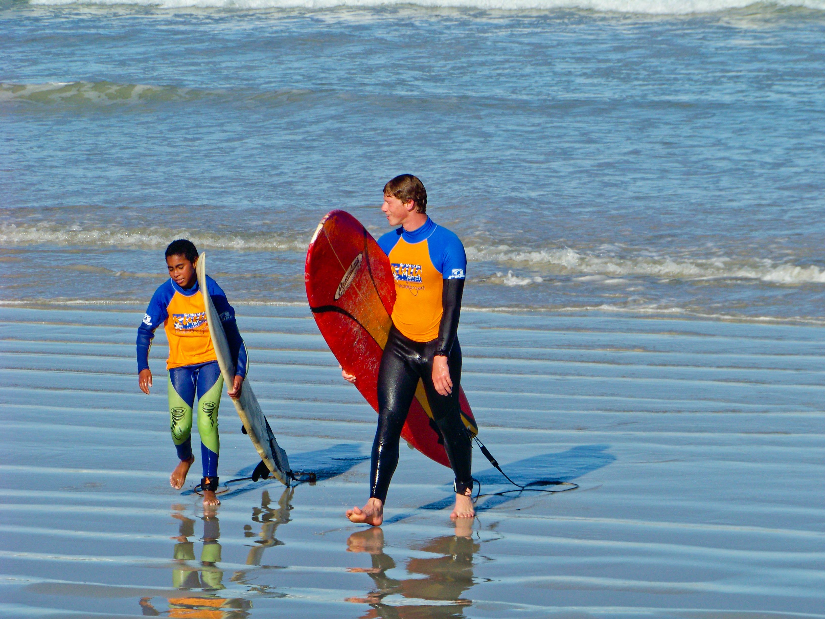 A Projects Abroad volunteer teaching children to surf in South Africa walks along the beach with a child A volunteer works closely with a child to teach surfing in South Africa